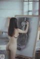 A nude woman standing in front of a drawing on a easel.